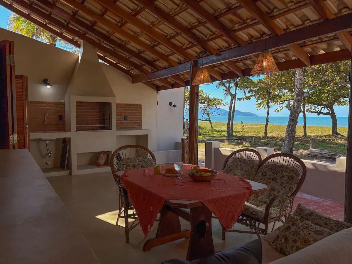 Varanda coberta da Casa Tohmé, com churrasqueira de tijolo, mesa posta e o mar visto entre as árvores